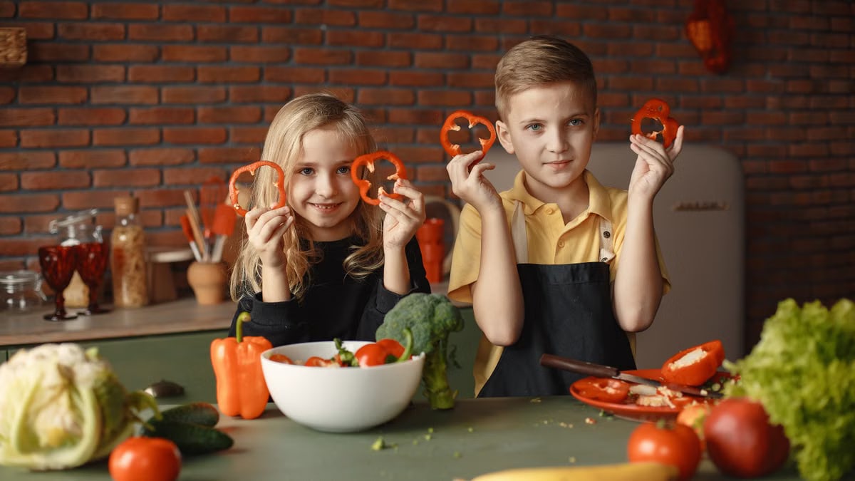 Two children holding capsicum slices while preparing vegetables in a cozy kitchen setting.